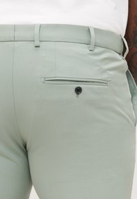 Κοντινό πλάνο ανοιχτοπράσινου παντελονιού με πίσω τσέπη welt κλειστή με μαύρο κουμπί, σε ένα άτομο που φοράει λευκό πουκάμισο.