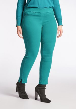 Femme portant un pantalon slim bleu sarcelle et des bottines noires à talons hauts, debout sur un fond blanc.