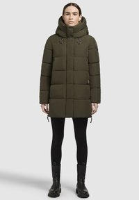 Palton pufos de culoare verde măsliniu, cu guler înalt, design matlasat și accente cu șnur. Purtați peste leggings negri și botine.