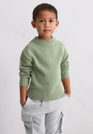 Groene gebreide trui met een geribbelde kraag, opgerolde mouwen en een aansluitende zoom, gecombineerd met lichtgrijze cargobroek met zijzakken.