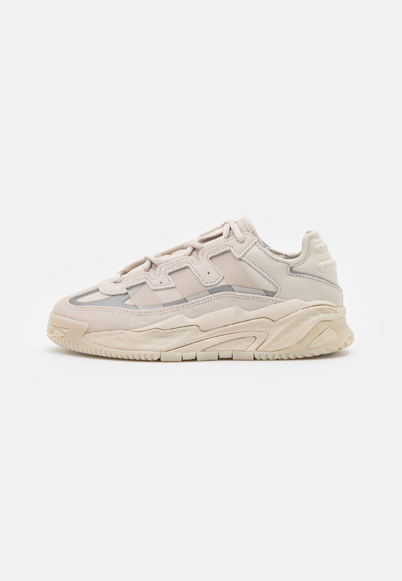 Zapatilla beige gruesa con una combinación de ante texturizado y cuero liso, que presenta paneles de malla y una suela de goma gruesa.