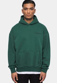 Donkergroene hoodie van fleece met een voorzak, ribgebreide boorden en een trekkoord in de capuchon. Heeft een subtiele geborduurde logo op de borst.