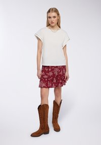 Top bianco a maniche corte, gonna rossa con volant floreale e stivali alti fino al ginocchio in suede marrone con ricami. Colori neutri, morbide texture, design a strati.