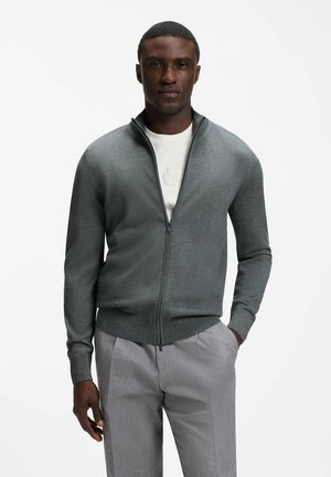 Hombre con un suéter gris con cremallera sobre una camisa blanca y pantalones gris claro, de pie con una mano en el bolsillo frente a un fondo liso.