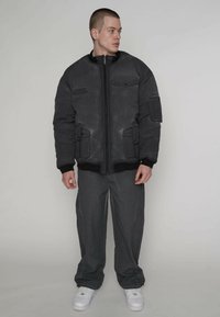 Zwarte gewatteerde jas met een ritssluiting aan de voorkant, zijzakken en geribbelde manchetten. Gestructureerde stof met lichte slijtage, gecombineerd met grijze broek en witte sneakers.