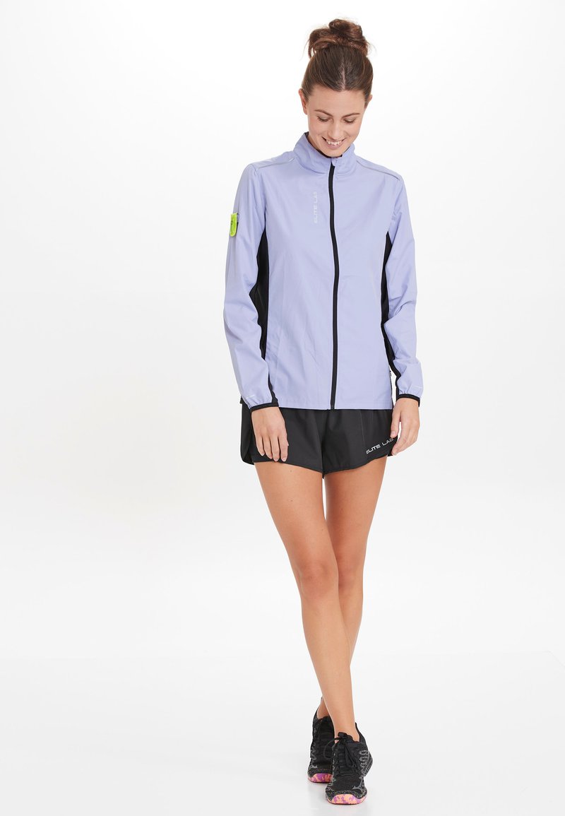 Veste de sport lavande clair avec des panneaux latéraux noirs, fermeture éclair intégrale, col montant et logo sur la poitrine. Associée à un short noir et des chaussures de course.