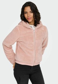 Svetlo roza puhast pulover z zadrgo, mehke teksture, z visokim ovratnikom in rebrastimi manšetami. Nošen preko belega topa s črnimi kavbojkami.