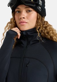 Black mid-layer jacket with a high collar, textured fabric, zippered front, and fitted sleeves. Features a small zippered pocket.