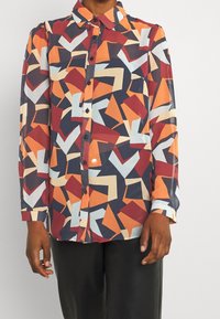 Blusa a maniche lunghe con bottoni in tessuto leggero, caratterizzata da un motivo geometrico nei colori rosso, arancione, blu navy e azzurro. Collo e polsini classici.