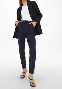 Marineblaue, schmal geschnittene Hose mit elastischem Bund und Kordelzug, kombiniert mit einem taillierten weißen Oberteil und einem schwarzen Blazer, abgerundet mit schwarzen Sandalen mit Riemchen.