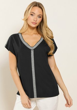 Lyshåret kvinde iført en sort kortærmet V-hals bluse med hvide stribede kanter, kombineret med hvide bukser, stående foran en ensfarvet baggrund.