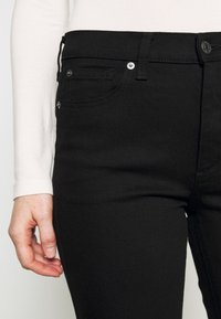 Jean en denim noir au design ajusté, avec des accessoires en métal argenté, des poches avant et une texture lisse. La taille comprend une fermeture à bouton.