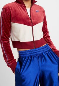 Röd och vit zip-up velourjacka med chevrondesign, som har blå detaljer och en Nike-logotyp. Matchad med blå satängshorts.