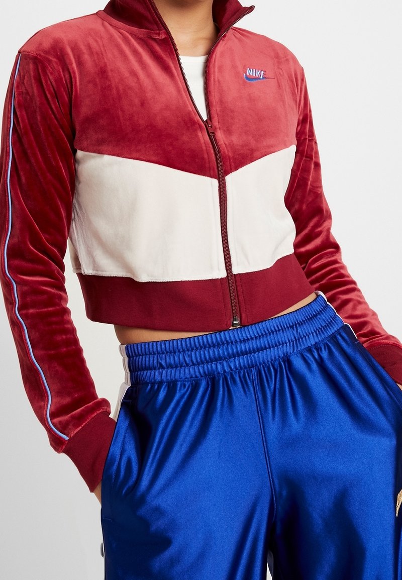Röd och vit zip-up velourjacka med chevrondesign, som har blå detaljer och en Nike-logotyp. Matchad med blå satängshorts.