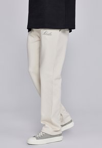 Beežid pehmetest kangast dressipüksid, mis on kitsas lõikega ning millel on vöökoha lähedal peen logo tikkimine, kantud koos hallide tossudega.