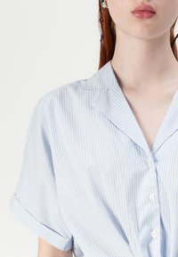 Camisa de botão azul claro com riscas brancas, com mangas curtas dobradas e colarinho pontudo. Tecido suave com uma textura lisa.