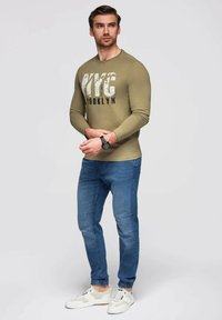 Camisa de manga larga verde oliva con estampado "NYC BROOKLYN", combinada con jeans ajustados azules y zapatillas blancas con acentos grises.