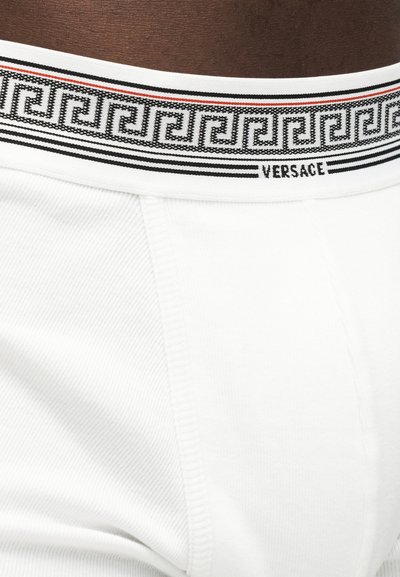 Sous-vêtements blancs Versace avec une ceinture noire et rouge à motifs, portés par une personne à la peau foncée.
