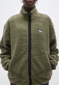 Grøn fleecejakke med høj krave, lynlås foran og sidelommer. Har en tekstureret overflade og et lille brandlogo på brystet.