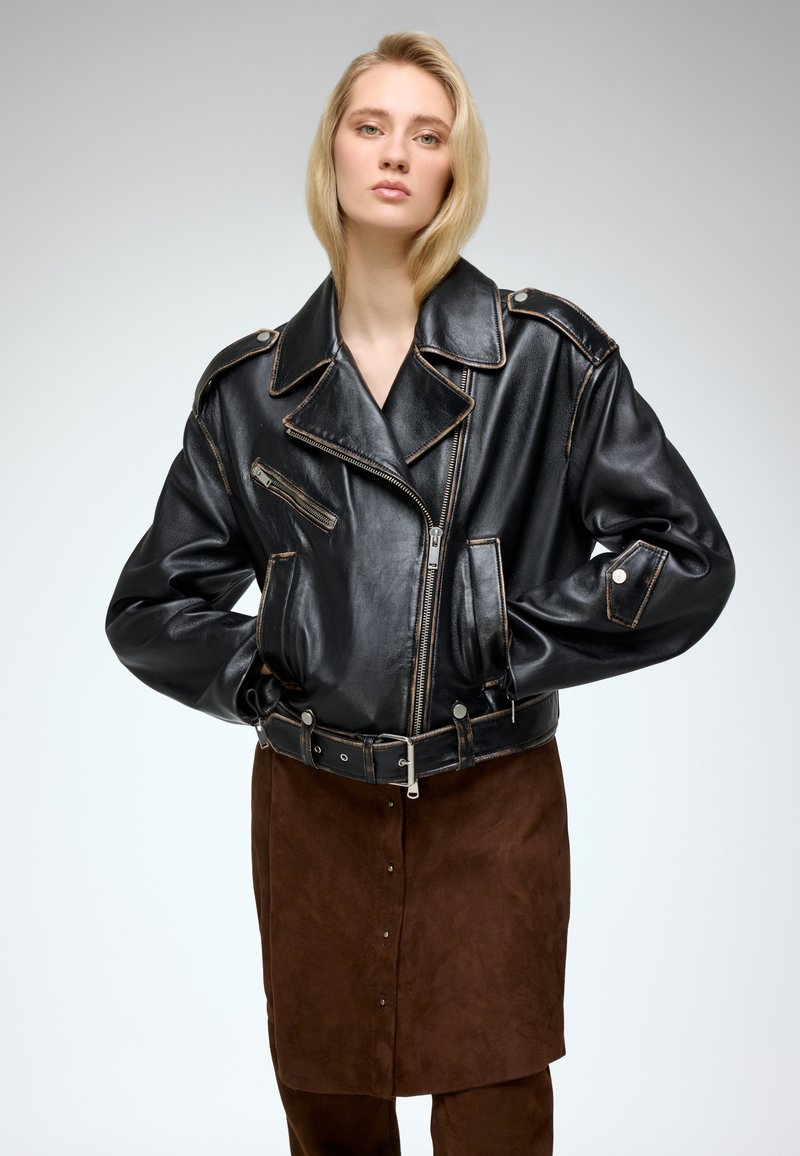 Black leather biker jacket with brown stitching, large lapels, zippered pockets, and a belted waist, paired with brown suede skirt.