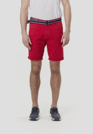 Røde bomuldsshorts med optrukket kant, forsynet med et navyblåt og hvidstribet bælte. Bæres med mørke sko og en hvid t-shirt.