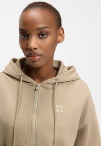 Frau mit kurzen Haaren, die einen beigefarbenen Kapuzenpullover mit Reißverschluss trägt, auf dessen Brust "HUGO" aufgestickt ist, blickt direkt nach vorne vor einem schlichten Hintergrund.