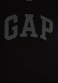 Melna kokvilnas T-kreklu ar lielu grafīta "GAP" logo, kas novietots centrā uz krūšu, ar apaļu apkakli un īsām piedurknēm.