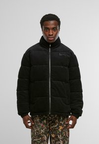 Mustast tumehari kangast pufferjakk kõrge kraega, ees tõmblukk ja tikkimismustriga. Vasakul küljel peen logo.