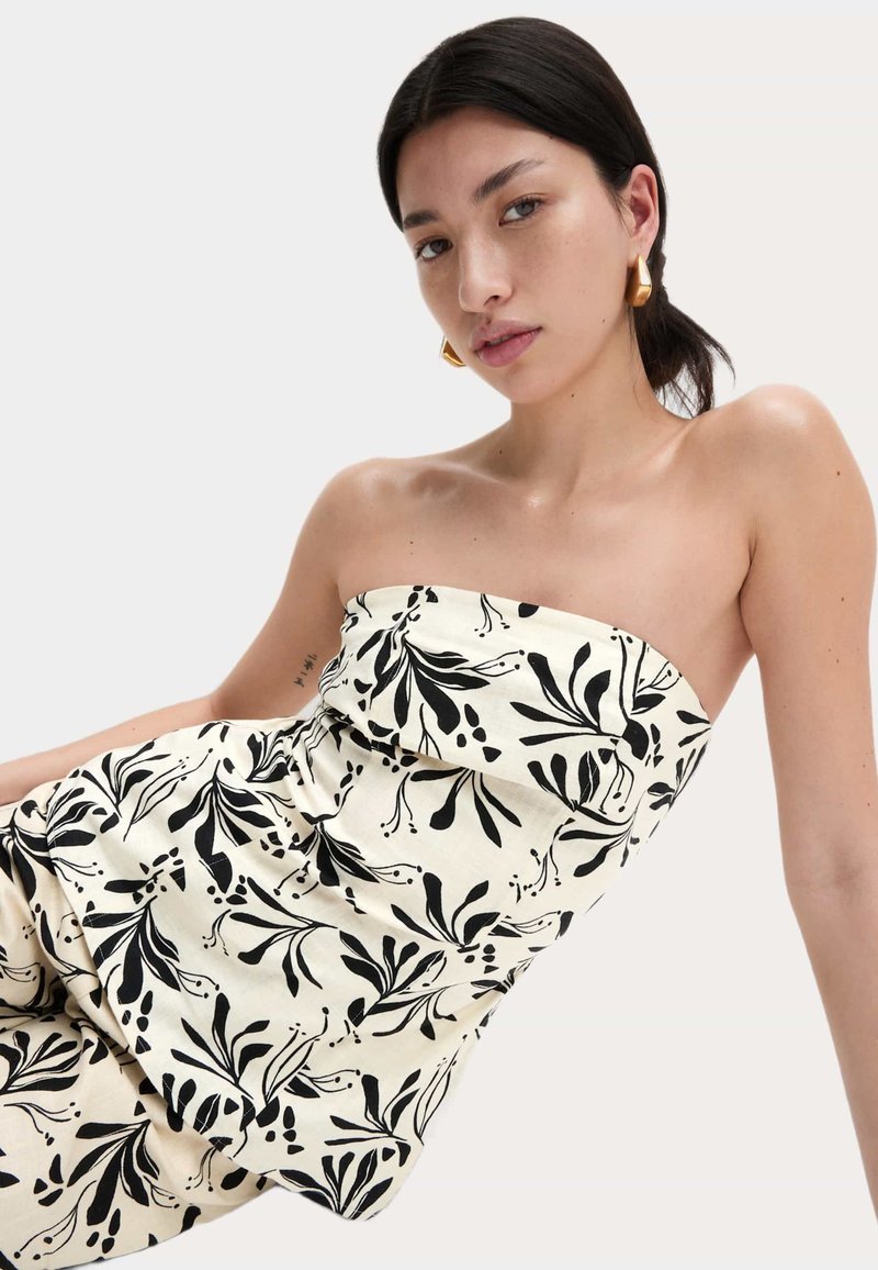 Jonge vrouw met donker haar in een zwart-wit bloemen strapless top en gouden hoepeloorbellen, poserend tegen een lichte, effen achtergrond.
