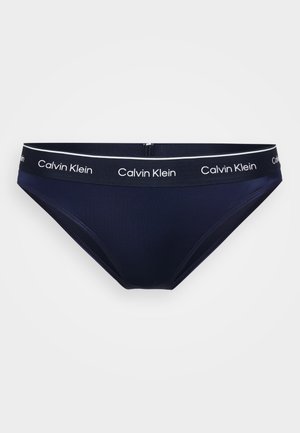 Bas de bikini bleu marine en tissu extensible, avec une ceinture en logo blanche portant le texte "Calvin Klein". Texture lisse et coupe classique.