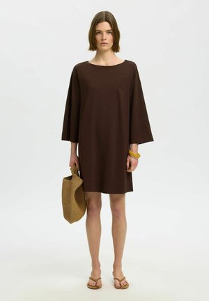 Mujer con un vestido suelto de color marrón oscuro que sostiene una bolsa de mano tejida, lleva sandalias color cuero y una pulsera amarilla gruesa, posando frente a un fondo liso.