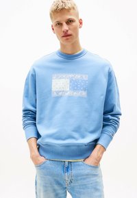 Sudadera azul claro con cuello redondo, manga larga y un gráfico estampado en blanco. Combinada con jeans azul desgastados.