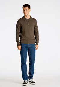 Brun stickad tröja med dragkedja i kragen, kombinerad med blå jeans. Jeansen har kontrasterande sömmar. Outfitten kompletteras med svarta sneakers.