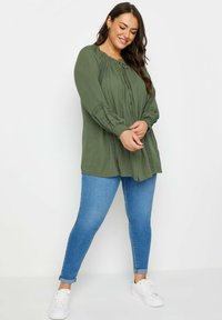 Olivgrön blus med plisserad design och knäppning framtill, i kombination med blå skinny jeans och vita sneakers. Långa ärmar samlade vid handlederna.