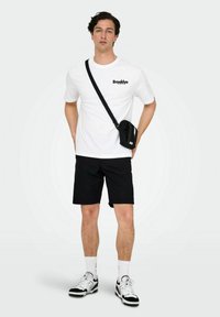 Hvid t-shirt med "Brooklyn" i sort tekst, sorte shorts, lille sort crossbody taske og sort-hvide sneakers.