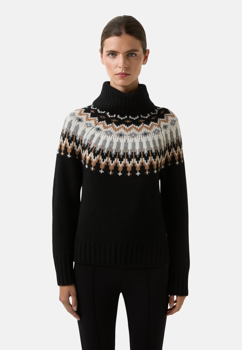 Vrouw in een zwarte coltrui met een bruin, wit en grijs geometrisch patroon op de borst en schouders, staande tegen een witte achtergrond.