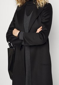 Cappotto nero con un design su misura, dotato di colletto e tasche. Indossato sopra un maglione a coste scuro, abbinato a una borsa nera.