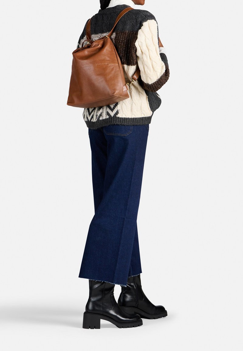 Zaino in pelle marrone con chiusura a cordoncino, indossato con un maglione a maglia grosso in grigio, crema e nero, abbinato a jeans a gamba larga blu e stivali neri.