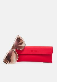 Un etui pentru ochelari de soare roșu din piele, cu o textură netedă, care conține ochelari de soare mari, de culoare maro deschis, translucizi, cu rame geometrice.