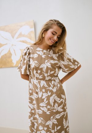 Kobieta uśmiechająca się z zamkniętymi oczami, ubrana w beżową sukienkę z białym wzorem liści, stojąca obok obrazu na ścianie z beżowymi i białymi liśćmi.