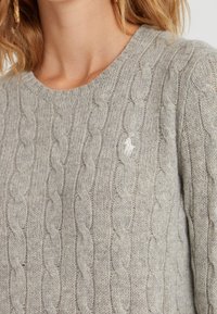 Harmaa palmikkoneulepusero, jossa pyöreä pääntie ja pieni brodeerattu logo. Neuleen pinta on pehmeä ja siinä on vaihtelevaa paksuutta olevia palmikoita.