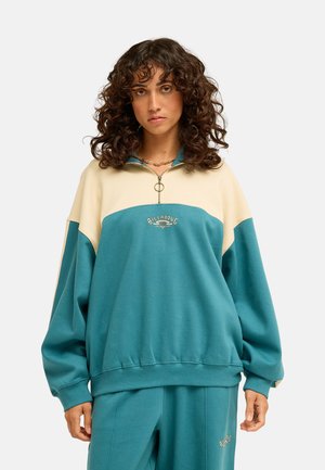 Donna con capelli ricci che indossa una felpa con zip Billabong turchese e crema e pantaloni turchese coordinati su uno sfondo chiaro e uniforme.