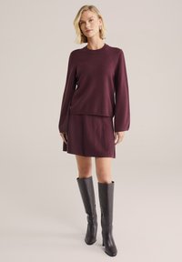 Bordeauxfarbener Strickpullover mit langen Ärmeln, kombiniert mit einem passenden Rock; dazu schwarze kniehohe Absatzstiefel mit glatter Textur.