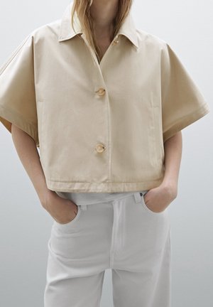 Vrouw draagt een beige kort mouwtjes overhemd met wijde mouwen en witte broek, handen in de zakken, staand tegen een effen achtergrond.