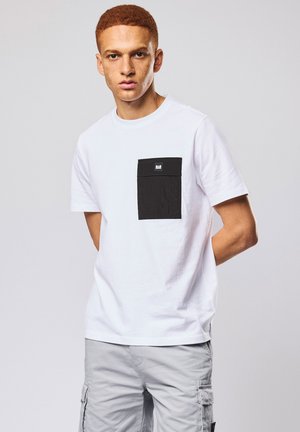 Jonge man met kort rood haar, gekleed in een wit T-shirt met een groot zwart borstzakje en lichtgrijze cargobroek.