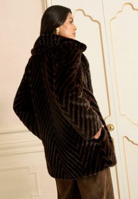 Žena s tmavohnedým pruhovaným umelým kožuchom, rukami v vreckách, stojaca bokom pri krémovej stene s dekoratívnym obložením.