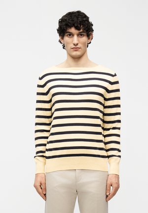 Fiatal férfi göndör hajjal, krémszínű és fekete vízszintes csíkos hosszú ujjú inget és bézs nadrágot visel, egyszínű háttér előtt áll.