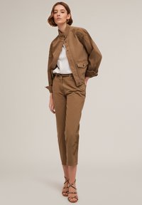 Bruine canvasjack met bloemmotieven op de schouders, gecombineerd met bijpassende aansluitende broek. Witte top eronder, sandalen met bandjes.