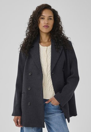Frau mit lockigem, dunklem Haar, die einen dunkelgrauen, doppelt geknöpften Mantel über einem cremefarbenen, grob gestrickten Pullover und blauen Jeans trägt, mit einer Hand in der Tasche.