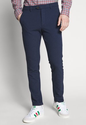 Mężczyzna ubrany w granatowe, zwężane spodnie, białe trampki z czerwonymi i zielonymi paskami oraz czerwoną kraciastą koszulę, z rękami w kieszeniach.